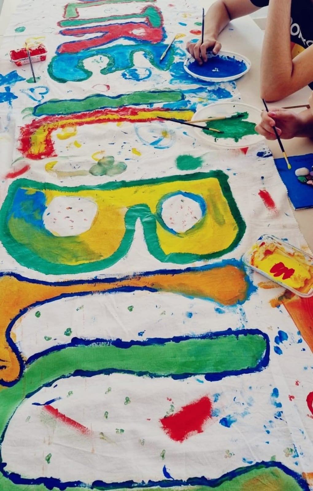 Mani e pennelli al lavoro su un grande foglio dipinto a colori vivaci, durante un laboratorio di pittura